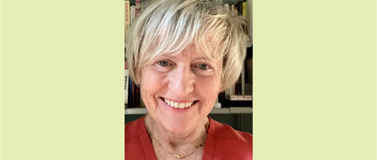Debbie Field smiling at the camera. She is a white woman with short blonde hair, wearing a red top. A shelf of books is in the background.