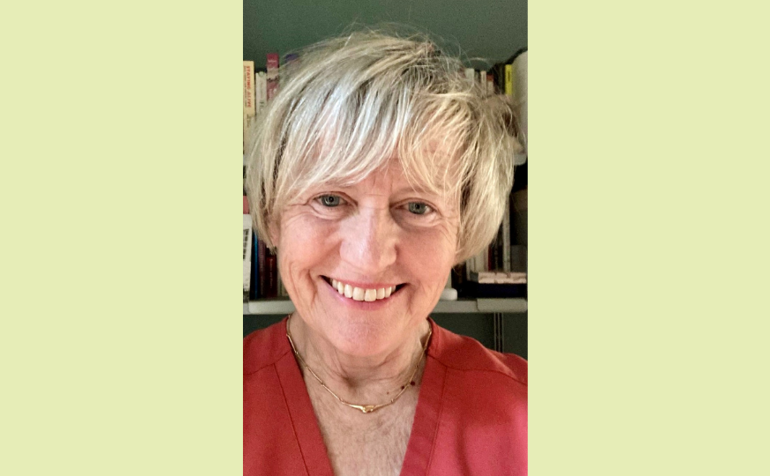Debbie Field smiling at the camera. She is a white woman with short blonde hair, wearing a red top. A shelf of books is in the background.